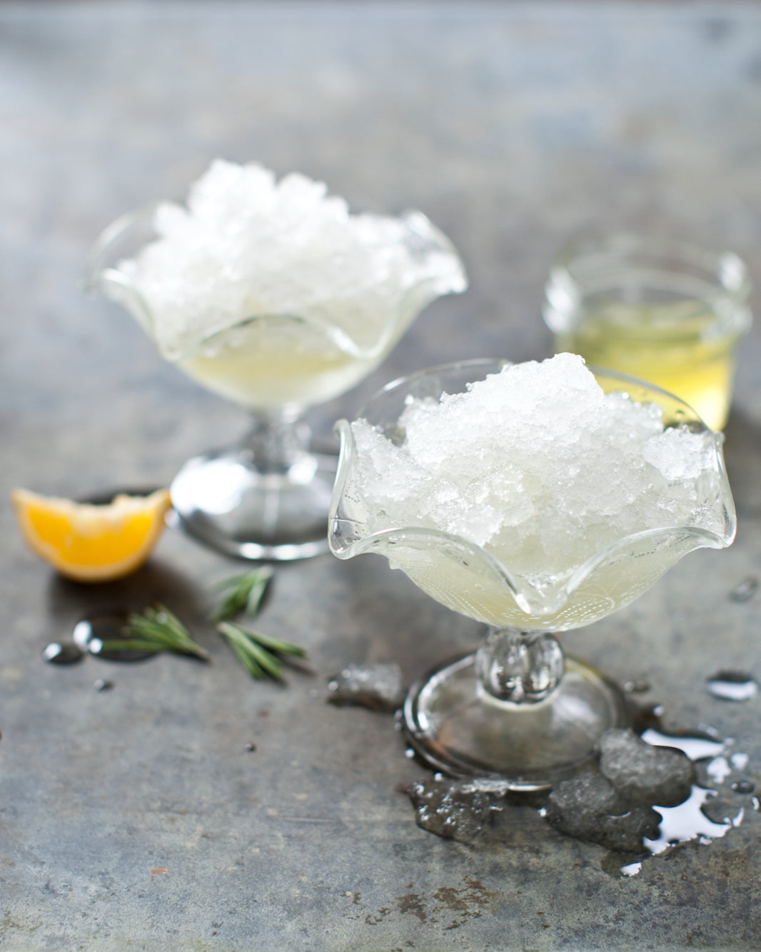 Two glass dessert bowls filled with shaved ice with lemon and rosemary in the background.