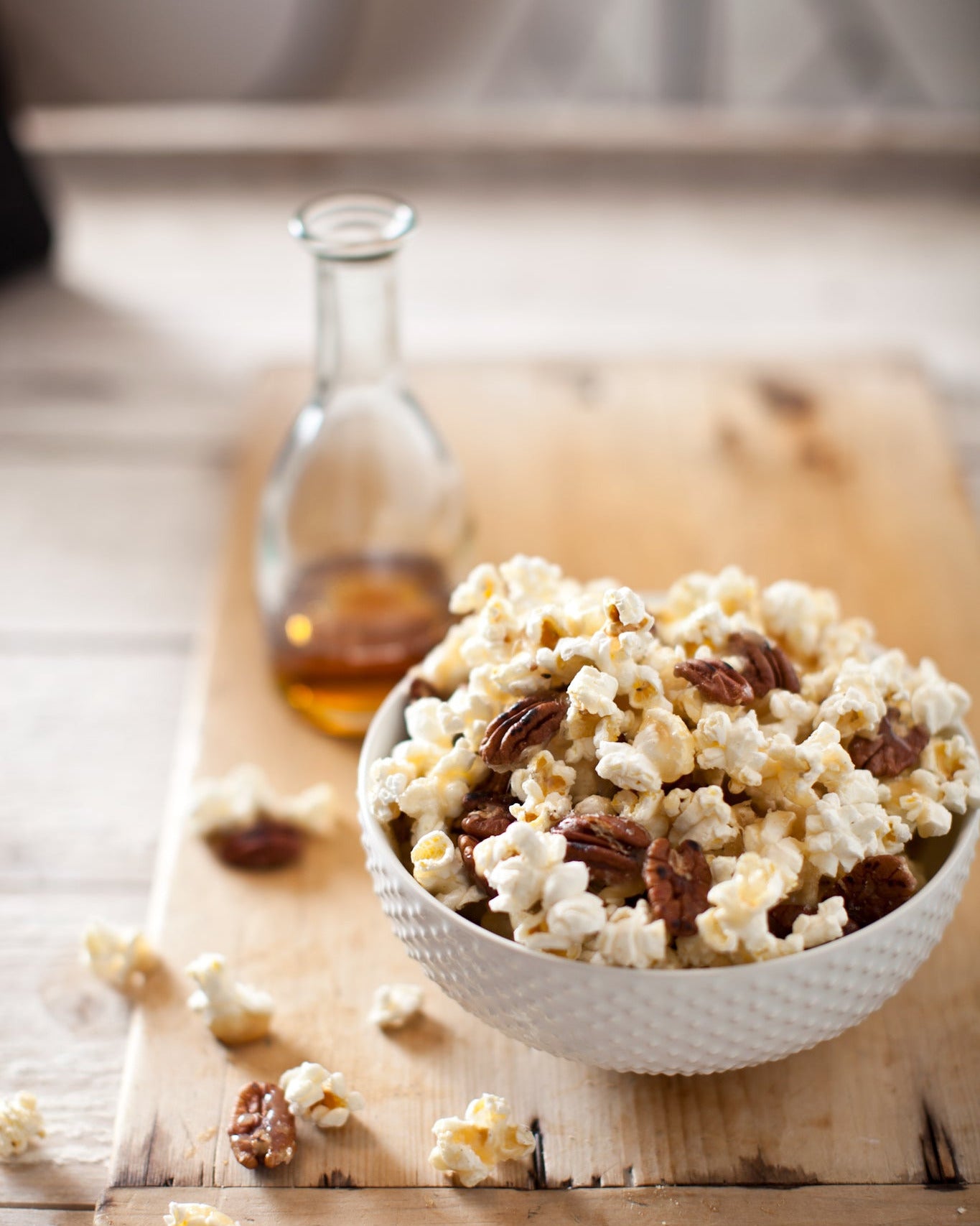 Bowl of maple caramel corn with pecans on a wooden board with a bottle of maple syrup.